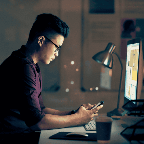 An entrepreneur sits at his desk looking at his computer and phone at that same time, overwhelmed by technical tasks
