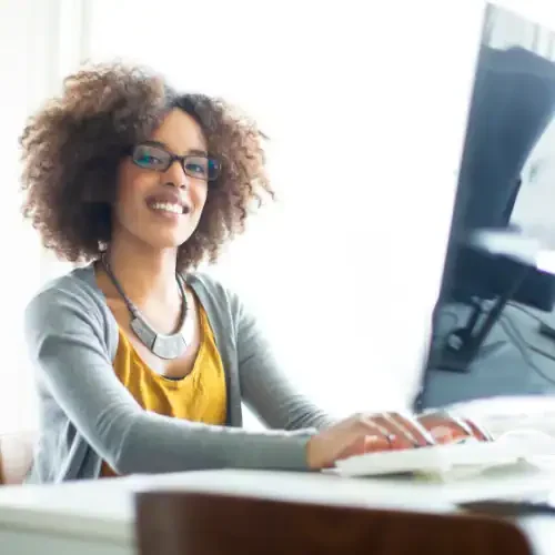 An entrepreneur smiles into the camera while working on her computer on work that she enjoys thanks to delegating tech tasks